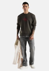 Sudadera verde oscuro con logo rojo de Calvin Klein, combinada con jeans grises y zapatillas blancas. Sosteniendo una chaqueta de color claro.