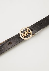 Zwarte synthetische leren riem met een gestructureerd krokodillendessin. Goudkleurige ronde gesp met "MK"-logo als het middelpunt.
