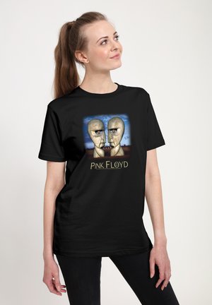 Femme portant un T-shirt noir Pink Floyd avec deux grandes têtes de pierre se faisant face sur un fond de ciel bleu.