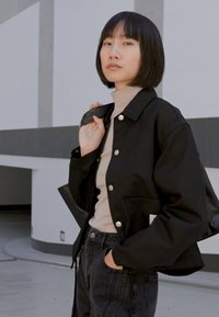Veste noire courte avec des boutons-pression argentés, portée par-dessus un col roulé beige clair, associée à un jean noir taille haute. Texture de tissu lisse.