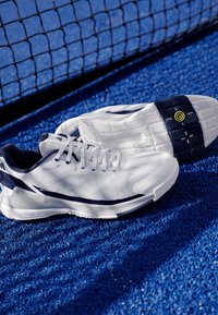Bílé sportovní boty s námornickými akcenty, strukturovaný síťový svršek a vzorovaná gumová podrážka, umístěné na modrém texturovaném povrchu.
