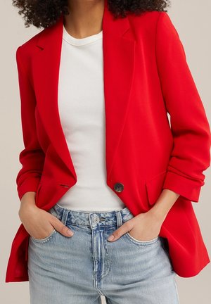 Osoba ubrana w jaskrawoczerwoną marynarkę na białej prążkowanej koszulce, z rękami w kieszeniach jasnoniebieskich dżinsów, stojąca na jednolitym tle.