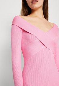 Donna che indossa un vestito di maglina rosa a coste con scollo off-shoulder e ampi lacci incrociati sul petto e in vita.
