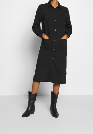 Abito nero a maniche lunghe con chiusura a bottoni, tasche sul petto e tasche laterali, indossato con stivali da cowboy neri. Tessuto morbido e design fino al ginocchio.