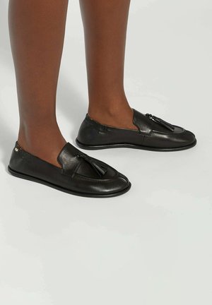 Jambes à la peau foncée portant des mocassins noirs en cuir à pampilles sur un sol gris clair, vues de côté et légèrement d'en haut.