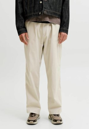 Mand iført beige løstsiddende bukser, beige og brune sneakers, en ternet skjorte og en mørk denimjakke mod en ensfarvet baggrund.