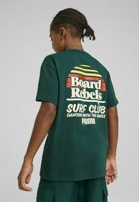 La camiseta de algodón verde presenta un estampado gráfico con franjas de atardecer en naranja y amarillo. El texto dice "Board Rebels Surf Club" y "PUMA."