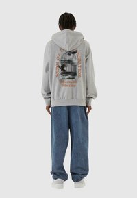 Grå hættetrøje med grafisk print, der viser et fuglebur og tekst på ryggen. Sammen med løse blå denimbukser og hvide sneakers.
