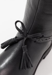 Botte en cuir noir dotée d'une tige texturée, d'accents décoratifs perforés et d'un détail de pompon noué à la cheville.