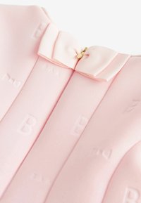 Różowa tkanina sukienki z wypukłymi literami, detalem kokardki przy dekolcie oraz złotym zamkiem błyskawicznym do zapinania. Gładka faktura i ukształtowany kształt.