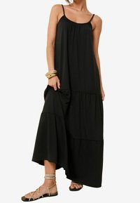 Svart maxiklänning med justerbara spaghettiband, lager-på-lager nederkant och avslappnad passform. Tillverkad av mjukt tyg och har volangdetaljer.