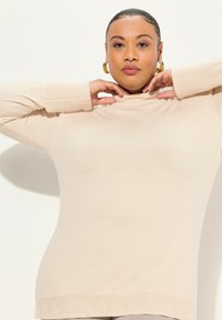Pullover beige a collo alto con una texture liscia, caratterizzato da polsini a coste e un delicato motivo triangolare all'orlo.