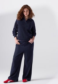 Dunkelblaues Sweatshirt mit Polokragen und lockeren Ärmeln, kombiniert mit locker sitzenden, dunkelblauen Jogginghosen und leuchtend roten Sportschuhen.