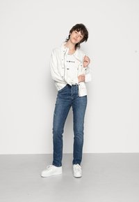Casaco de denim branco sobre uma camisa branca com uma estampa preta, combinado com jeans azuis e ténis brancos. Design simples com um aspeto casual.
