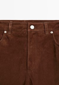 Brązowe zamszowe spodnie o gładkiej fakturze, z zapięciem na guziki i metalowymi akcentami. Podwójnie przeszywane szwy wzdłuż pasa i kieszeni.