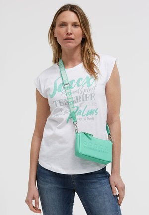 Woman wearing a white graphic tee and blue jeans, carrying a mint green crossbody bag with a wide strap across her chest.