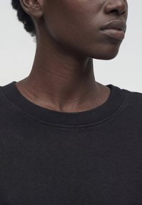 Κοντινό πλάνο του κάτω μέρους του προσώπου και του λαιμού ενός ατόμου που φοράει μαύρο μπλουζάκι με στρογγυλή λαιμόκοψη, με φόντο έναν απλό ανοιχτόχρωμο φόντο.