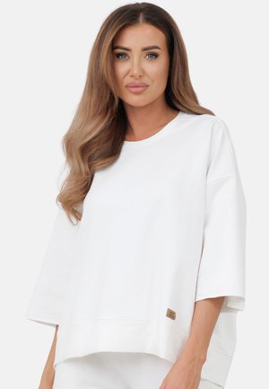 Kobieta z długimi brązowymi włosami, ubrana w luźny biały krótki top z krótkim rękawem, stojąca na tle jednolitego jasnego tła, zwrócona przodem.