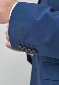 Ärmel eines blauen Anzugsjackets mit fünf braunen Knöpfen und blauer Naht. Der Stoff wirkt glatt mit einer subtilen Textur.