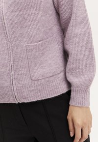 Lila stickad cardigan med ribbad textur, långa ärmar och en framficka. Mjuk tyg med avslappnad passform och synliga sömsdetaljer.