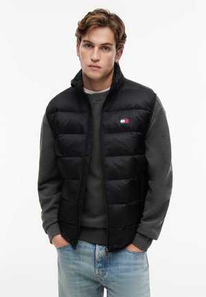 Giovane uomo con capelli mossi indossa un gilet imbottito nero sopra un maglione grigio scuro e jeans azzurri, in piedi con le mani nelle tasche davanti a uno sfondo bianco.