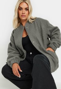 Grå fleecejacka med krage, tryckknappar och stora ärmar, kombinerad med en svart topp och mörka byxor. Mjuk struktur.