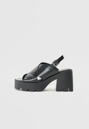 Mustat lakatut nahkaiset slingback-alustasaappaat paksulla korolla, joissa on ristivöiden muotoilu ja teksturoitu pohja pidon varmistamiseksi.