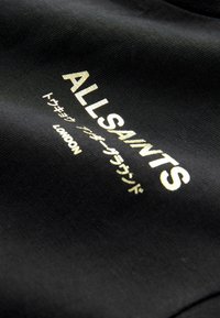 Melnā T-kreklu ar zelta burtiem, kas lasa "ALLSAINTS" un papildu tekstu japāņu valodā, ar gludu sintētisku tekstūru.