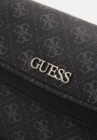 Černá syntetická peněženka s jemným embosovaným vzorem. Stříbrné logo "GUESS" umístěné na prominentním místě. Hladký povrch s kontrastním lemem.