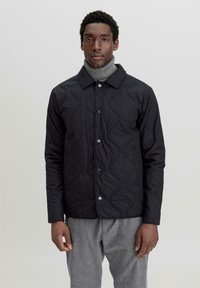 Schwarze gesteppte Jacke mit glatter Textur, runder Kragen und Druckknopfverschluss; getragen über einem grauen Rollkragenpullover.