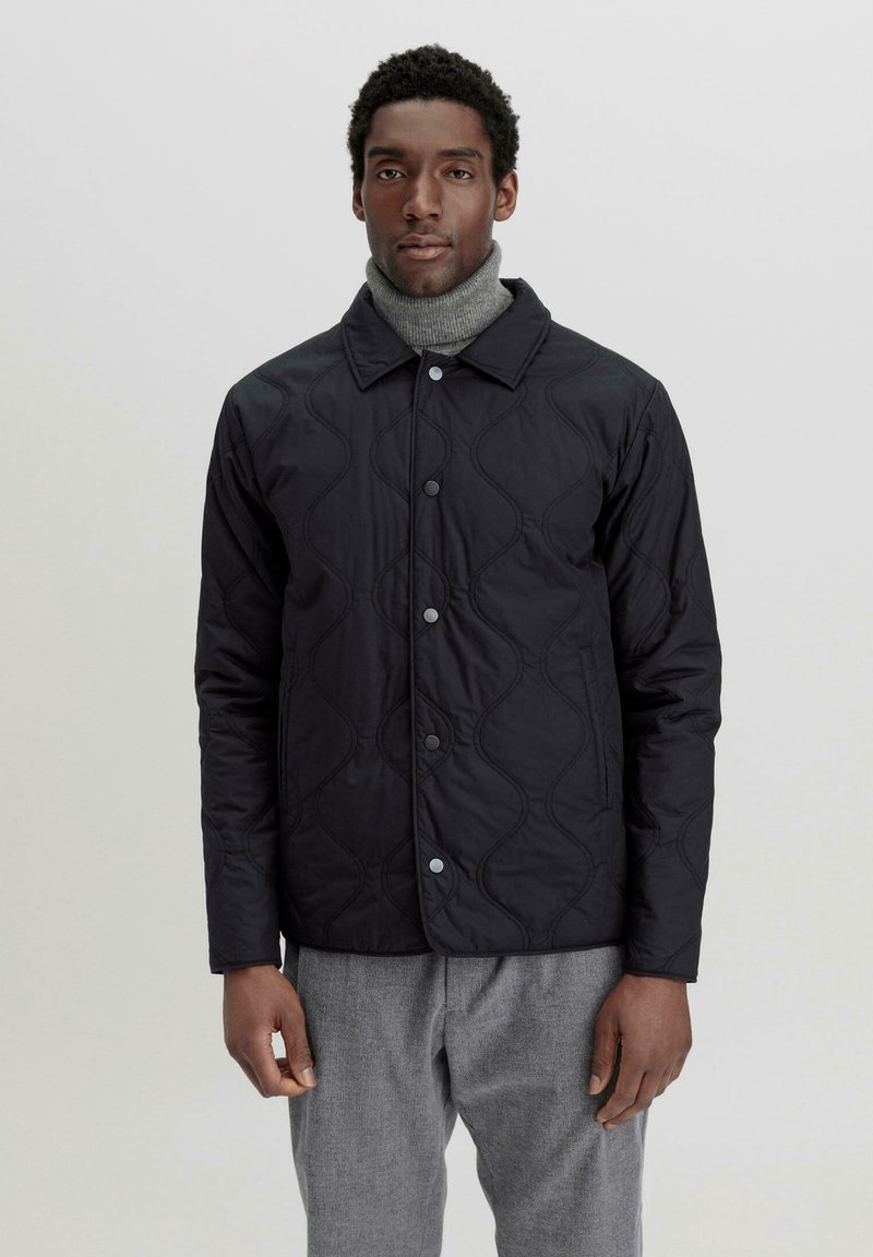 Schwarze gesteppte Jacke mit glatter Textur, runder Kragen und Druckknopfverschluss; getragen über einem grauen Rollkragenpullover.