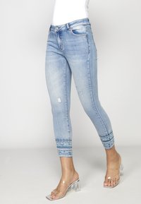 Jean skinny bleu clair avec une légère usure et des bords effilochés à motifs, porté avec des sandales à talons hauts transparentes sur un fond blanc.