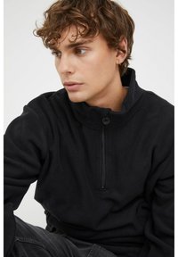 Pull en polaire noir avec un col montant et un demi-zip. Texture douce et coupe décontractée. Logo discret sur la poitrine.