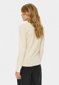 Gara piedurkņu vieglais džemperis gaisīgi beige krāsā ar viļņotu apakšmalu. Audums izskatās mīksts, ar gludu tekstūru.