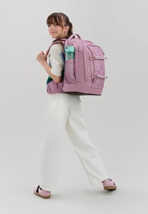 Young girl wearing a large pink backpack with a water bottle in side pocket, looking over shoulder, standing against a plain background.