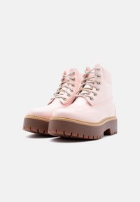 Γυαλιστερά παστέλ ροζ πλατφόρμα μποτάκια με κορδόνια, χοντρές καφέ σόλες και ασημί κρίκους, τοποθετημένα πάνω σε απλό ανοιχτό φόντο.