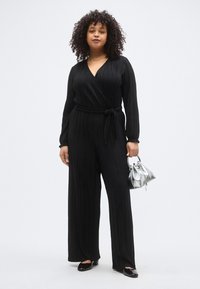 Juodas klostuotas kombinezonas su apvijimo dizainu, ilgomis rankovėmis, surištu liemeniu ir platėjančiomis kelnėmis. Derinamas su maža sidabrine rankine ir juodais balerinais.