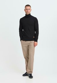 Hombre de pie con suéter negro de cuello alto, pantalones beiges y zapatillas negras contra un fondo claro.