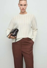 Pull en tricot crème avec un motif en chevrons, associé à un pantalon marron cintré, tenant un petit sac à main noir avec fermeture à cordon.