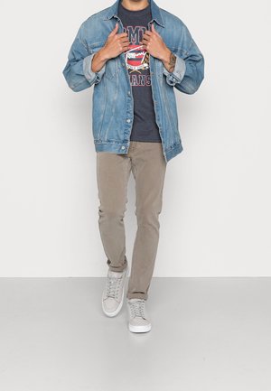 Homme ajustant une veste en denim bleu clair sur un t-shirt graphique sombre, un pantalon beige et des baskets gris clair sur fond blanc.