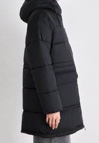 Osoba w długiej, czarnej, ocieplanej kurtce zimowej z kapturem, stojąca bokiem na jednolitym jasnym tle.