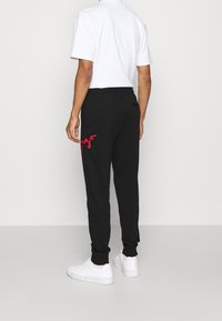 Schwarze Jogginghosen mit schmal zulaufenden Beinen, die ein rotes gesticktes Logo am linken Bein haben. Hergestellt aus einem weichen Stoff mit einem elastischen Bund.