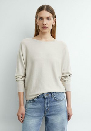 Jonge vrouw met stijl lichtbruin haar draagt een lichtbeige trui en blauwe spijkerbroek, staand tegen een effen witte achtergrond.