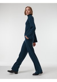 Suéter de punto en color verde azulado oscuro con cuello de tortuga y mangas largas, combinado con pantalones de punto acampanados a juego. El modelo presenta bolsillos laterales y zapatos planos.