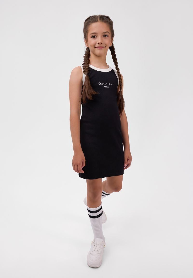 Jeune fille aux cheveux tressés portant une robe noire sans manches, des baskets blanches et des chaussettes blanches hautes avec des rayures noires.