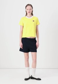 Gul cropped t-shirt med hvid krave og logo-patch, parret med mørkeblå shorts. Modellen bærer hvide strømper og sorte flade sko.