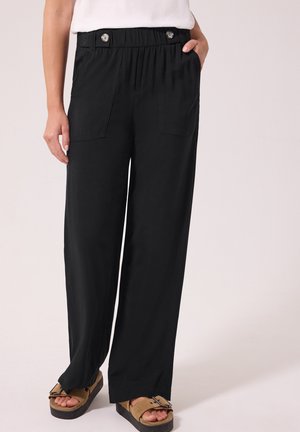 Personne portant un pantalon noir à jambes larges avec taille élastique et boutons, associé à des sandales marron à boucle et un haut blanc.
