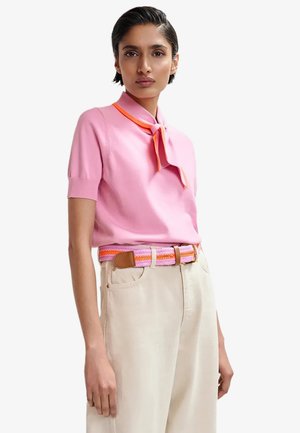 Donna che indossa una maglietta rosa a maniche corte con una cravatta bordata di arancione, pantaloni beige e una cintura intrecciata rosa e arancione.