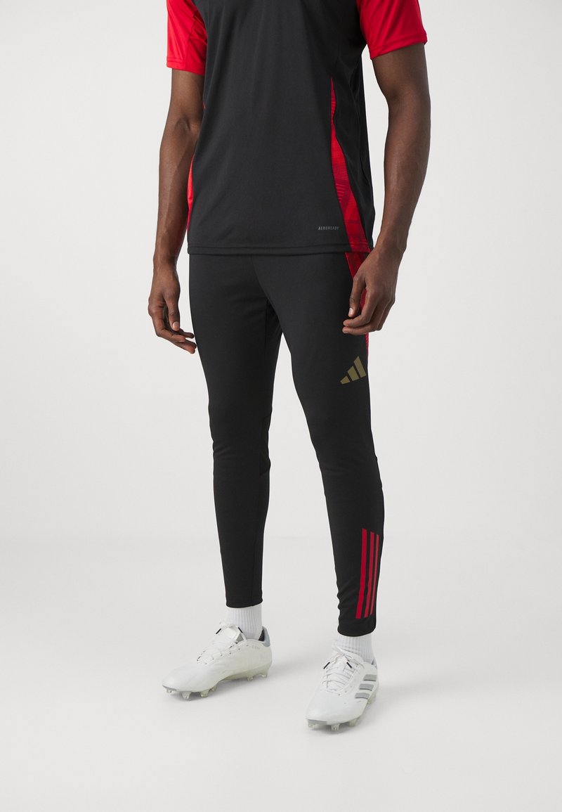 Schwarzes Sporthemd mit roten Ärmeln, schwarze enganliegende Leggings mit goldenem Logo und roten Streifen, kombiniert mit weißen Fußballschuhen.