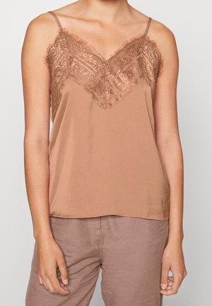 Femme portant un top en soie marron clair sans manches avec bordure en dentelle et un pantalon marron clair assorti, debout devant un fond uni.